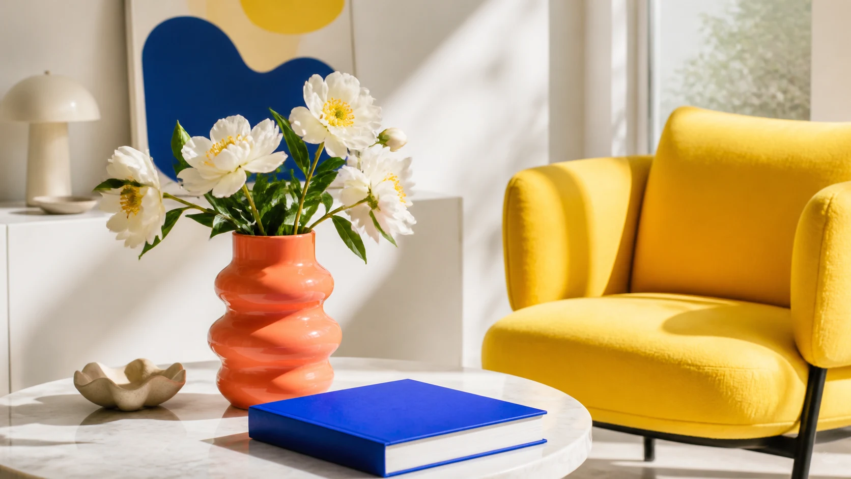 A coral vase, blue book, yellow chair, and white flowers in a sunlit modern room