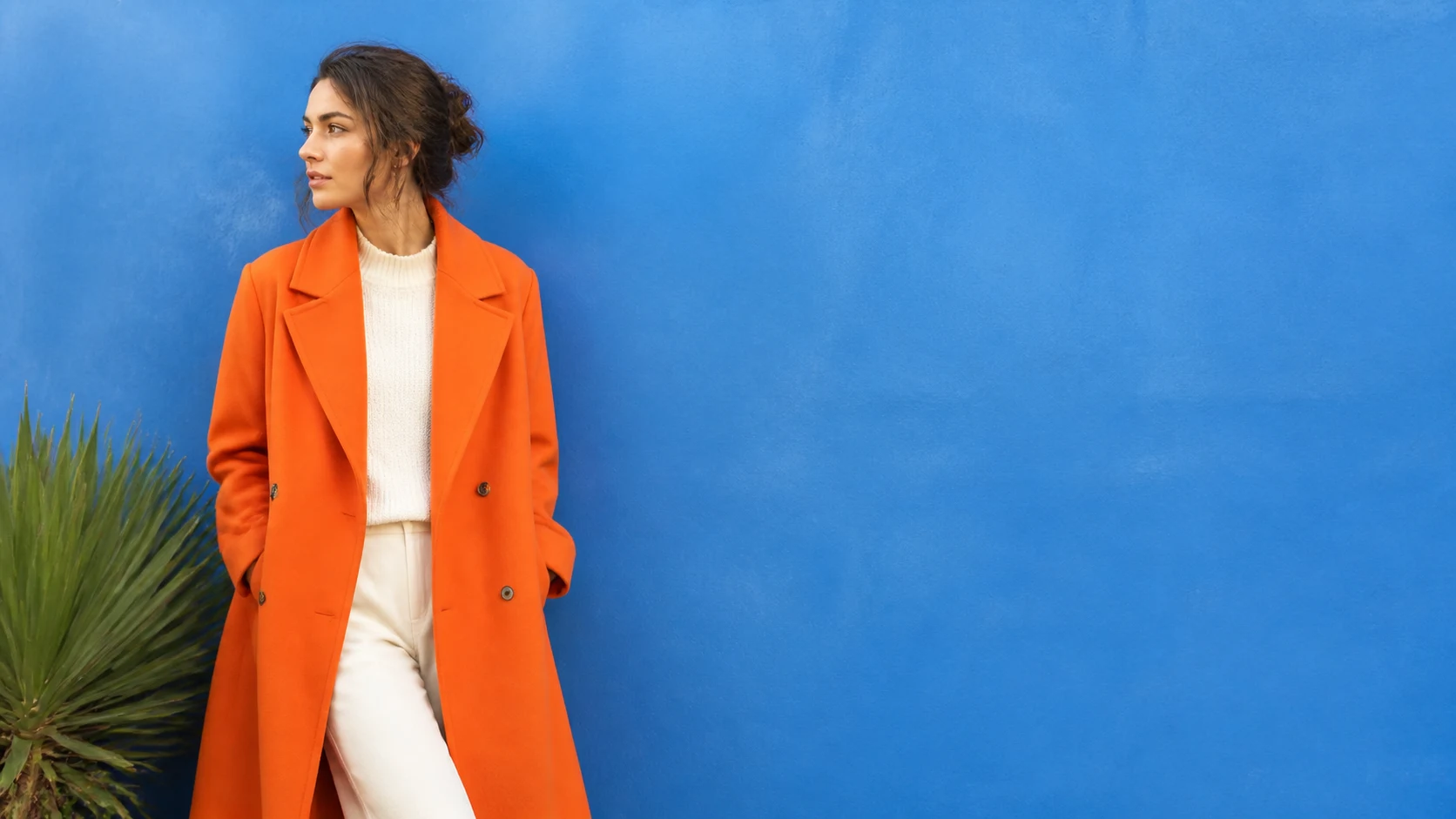 A fashion editorial portrait in an orange coat beside a saturated blue wall and green plant